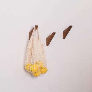 May include: Three wooden wall hooks in a triangular design are mounted on a white wall. A beige mesh bag filled with yellow lemons hangs from one of the hooks. The hooks are made of dark wood.
