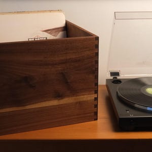 May include: A dark brown wooden storage crate with visible finger joints at the corners, containing a vinyl record. Next to the crate is a black turntable with a clear plastic dust cover, featuring a circular platter and a central spindle.