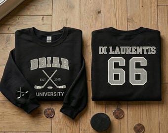 May include: Two black sweatshirts with hockey-themed designs are displayed on a wooden surface. One sweatshirt features "Briar University" with crossed hockey sticks. The other displays "Di Laurentis 66". A hockey puck and coins are also present.