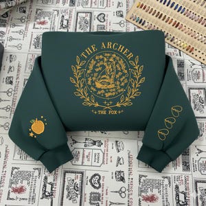 May include: Dark green sweatshirt with gold embroidery. The front features a circular design with a fox and the text "THE ARCHER" and "THE FOX". The left sleeve has an embroidered apple, and the right sleeve has three heart shapes.