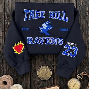 K&ouml;nnte beinhalten: Marineblaues Sweatshirt mit dem Text "TREE HILL RAVENS" in Wei&szlig; und Blau. Es zeigt eine blaue Raben-Grafik, die Nummer 23 und einen Aufn&auml;her mit Herz und Flammen. Ein Sweatshirt im klassischen Stil.