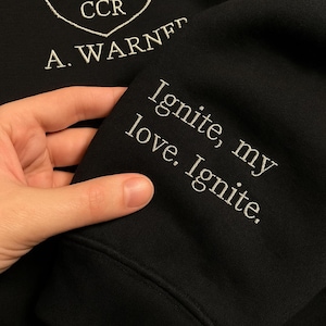 May include: Black sweatshirt with white embroidered text: "Ignite, my love. Ignite." The sleeve also features the embroidered name "A. WARNER" and the letters "CCR" inside a heart-shaped design.