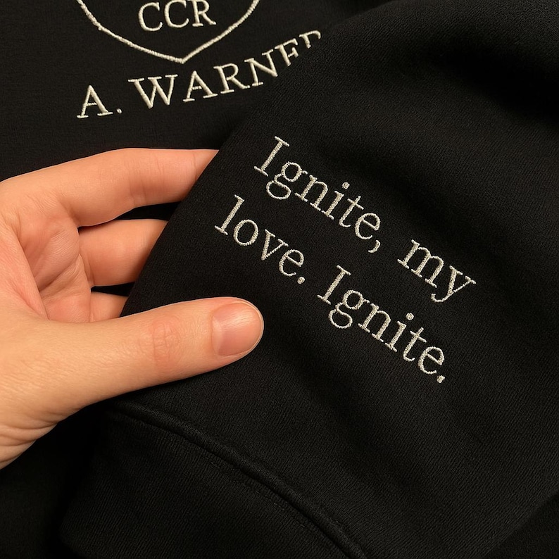 May include: Black sweatshirt with white embroidered text: "Ignite, my love. Ignite." The sleeve also features the embroidered name "A. WARNER" and the letters "CCR" inside a heart-shaped design.