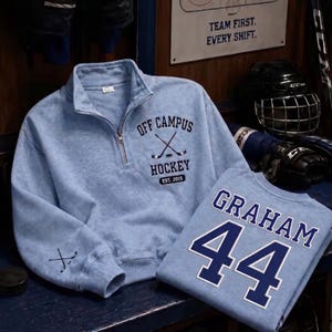 Op de afbeelding: Lichtblauwe hockey-thema sweatshirts. Eén heeft "OFF CAMPUS HOCKEY EST. 2015" met gekruiste hockeysticks. De andere heeft "GRAHAM 44" erop. Beide liggen op een donkerblauwe ondergrond.