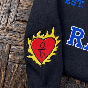 K&ouml;nnte beinhalten: Schwarzes Sweatshirt mit einem herzf&ouml;rmigen Aufn&auml;her in Rot mit gelbem Flammendesign. Das Herz enth&auml;lt die Buchstaben "c" und "b". Das Wort "EST." ist blau und der Buchstabe "R" ist wei&szlig;.