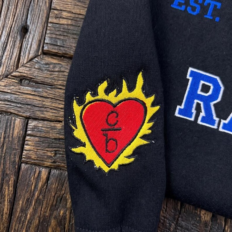 K&ouml;nnte beinhalten: Schwarzes Sweatshirt mit einem herzf&ouml;rmigen Aufn&auml;her in Rot mit gelbem Flammendesign. Das Herz enth&auml;lt die Buchstaben "c" und "b". Das Wort "EST." ist blau und der Buchstabe "R" ist wei&szlig;.