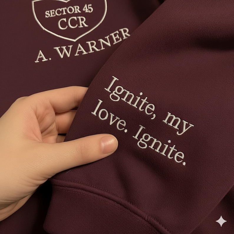 May include: A maroon sweatshirt with white embroidered text and a shield logo. The sleeve features the text "Ignite, my love. Ignite." The chest displays "Sector 45 CCR" and "A. Warner" in white.