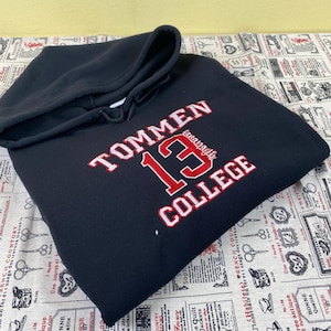 Peut inclure: Un sweat-shirt à capuche bleu marine avec le texte "TOMMEN 13 COLLEGE" en rouge et blanc. Le sweat-shirt a une capuche à cordon et une poche kangourou. Le vêtement est fait d'une matière douce et confortable.