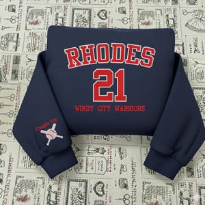 May include: A navy blue sweatshirt featuring the name "RHODES" and the number "21" in red. Below the number, the text reads "WINDY CITY WARRIORS" in red. The left sleeve displays a baseball graphic and the words "Caught Up".