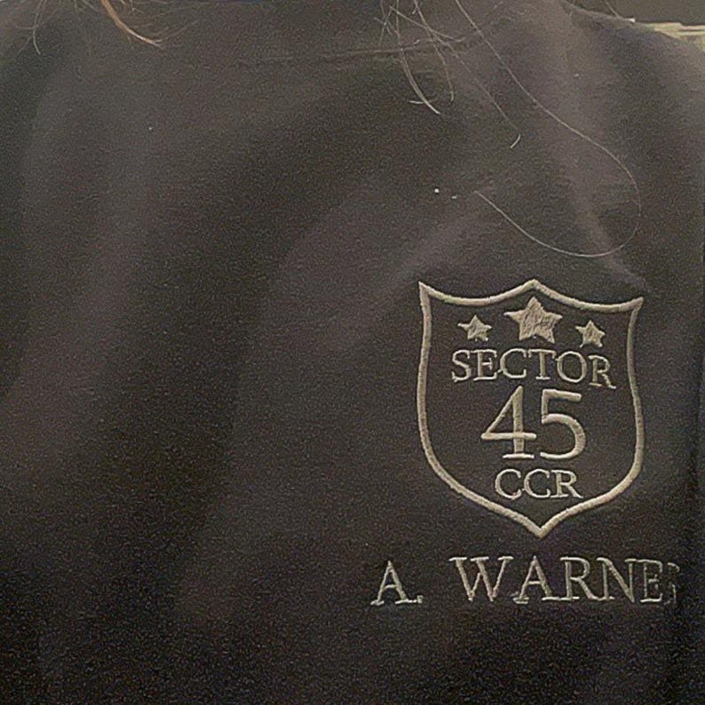 May include: Dark navy sweatshirt with an embroidered crest. The crest displays the text "SECTOR 45 CCR" and the name "A. WARNER" beneath. The crest is shield-shaped with star details.