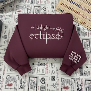 May include: A maroon sweatshirt with the text "the twilight saga eclipse" in white on the front. The right sleeve has the text "look after my heart, I've left it with you" in white. The sweatshirt is made of a soft material.