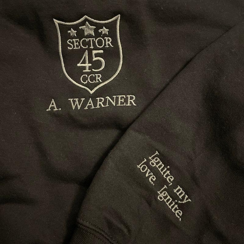 May include: Black sweatshirt with white embroidered text and design. The design includes a shield with "SECTOR 45 CCR" and the name "A. WARNER". The sleeve features the text "Ignite, my love. Ignite."