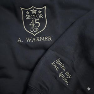 May include: Dark navy sweatshirt with embroidered details. The front displays a shield design with "SECTOR 45 CCR" and "A. WARNER". The sleeve is embroidered with the words "Ignite, my love. Ignite."