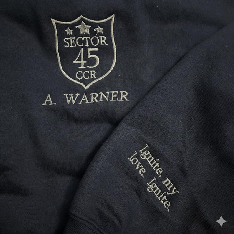 May include: Dark navy sweatshirt with embroidered details. The front displays a shield design with "SECTOR 45 CCR" and "A. WARNER". The sleeve is embroidered with the words "Ignite, my love. Ignite."