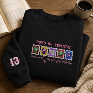 May include: A black crewneck sweatshirt featuring the text BOYS OF TOMMEN in pink embroidery. The design includes five colorful postage stamp graphics of sports jerseys and signatures. The left sleeve displays the number 13 in pink and white.