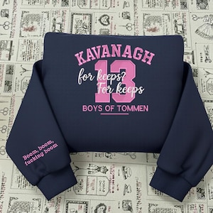 Puede incluir: Sudadera azul marino con texto rosa: "KAVANAGH 13 for keeps? For keeps BOYS OF TOMMEN." La manga izquierda muestra "Boom, boom, fucking boom" en rosa. La prenda está doblada, mostrando el diseño.