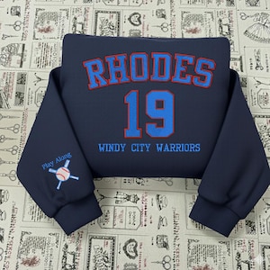 May include: A navy blue sweatshirt featuring the word "RHODES" in red and blue lettering above the number "19". The words "WINDY CITY WARRIORS" are below the number. The left sleeve has a baseball graphic with the words "Play Along".