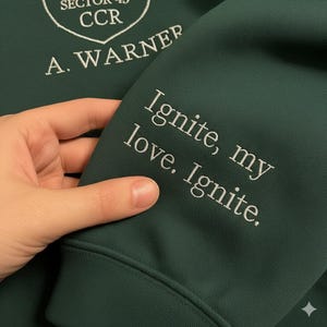 May include: Dark green hoodie with white embroidered text on the sleeve that reads "Ignite, my love. Ignite." The upper left has white embroidered text that reads "Sector 45 CCR" and "A. Warner".