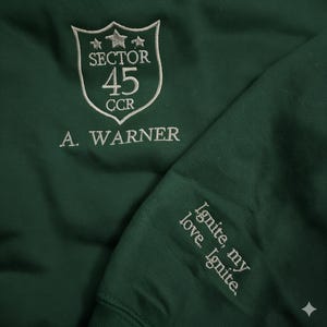 May include: Dark green sweatshirt with white embroidered text and design. The design includes a shield with "SECTOR 45 CCR" and "A. WARNER". The sleeve features the text "Ignite, my love. Ignite."