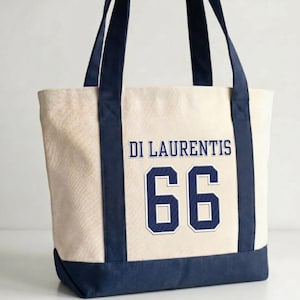 Puede incluir: Bolso tote color crema con detalles en azul marino. La bolsa presenta el texto "DI LAURENTIS" sobre el número "66" en azul marino. Las asas y la parte inferior de la bolsa son de color azul marino.