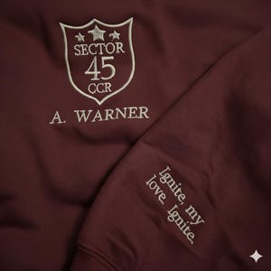 May include: A maroon sweatshirt with white embroidered text and graphics. The front features a shield design with "SECTOR 45 CCR" and "A. WARNER." The sleeve displays "Ignite, my love. Ignite."