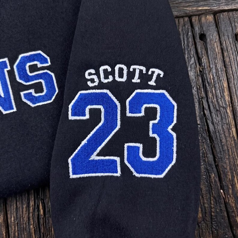 K&ouml;nnte beinhalten: Nahaufnahme eines marineblauen Sweatshirt-&Auml;rmels. Der &Auml;rmel zeigt den Namen "SCOTT" und die Nummer "23" in Wei&szlig; mit blauem Umriss. Der Buchstabe "S" ist ebenfalls sichtbar. Der Hintergrund ist eine Holzoberfl&auml;che.