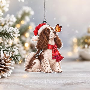 May include: A hanging ornament featuring a brown and white spaniel dog wearing a red Santa hat and a red scarf with white snowflake patterns. A monarch butterfly rests on the dog's nose. The name Charlie is printed in white script on the dog's chest.