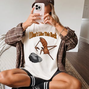 May include: An ivory t-shirt featuring a brown insect illustration and the text "I Don't Like This Rock" in brown letters above a grey rock. The shirt is worn with a brown plaid button-down shirt, a brown baseball cap, and black shorts.