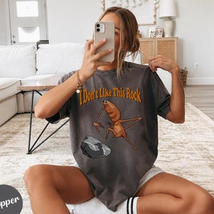 May include: A charcoal gray short-sleeve t-shirt featuring a graphic of a brown insect-like creature standing near a gray rock. The text "I Don't Like This Rock" is printed in orange above the creature. The shirt has a loose, relaxed fit.