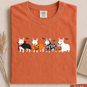 May include: A burnt orange crew neck t-shirt featuring a graphic of four white Bull Terrier dogs in Halloween costumes, including a vampire, a pumpkin, a skeleton, and a ghost, with small black bats printed above the dogs and a Comfort Colors label.