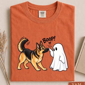 May include: A burnt orange crew neck t-shirt featuring a graphic of a German Shepherd dog and a white ghost. The ghost is touching the dog's nose with the text BOOP! and a small heart printed above them. The shirt has a Comfort Colors brand label.
