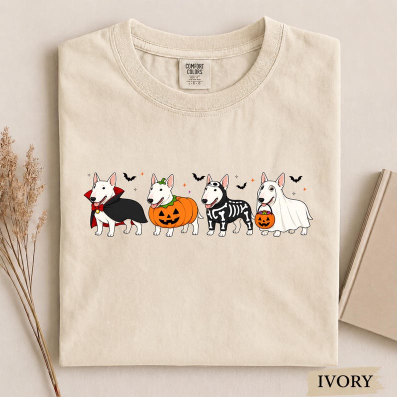 Puede incluir: Una camiseta de color marfil con un gr&aacute;fico de cuatro perros Bull Terrier blancos disfrazados de Halloween: un vampiro con capa, una calabaza, un traje de esqueleto y un fantasma con un cubo de calabaza, decorado con murci&eacute;lagos y estrellas.