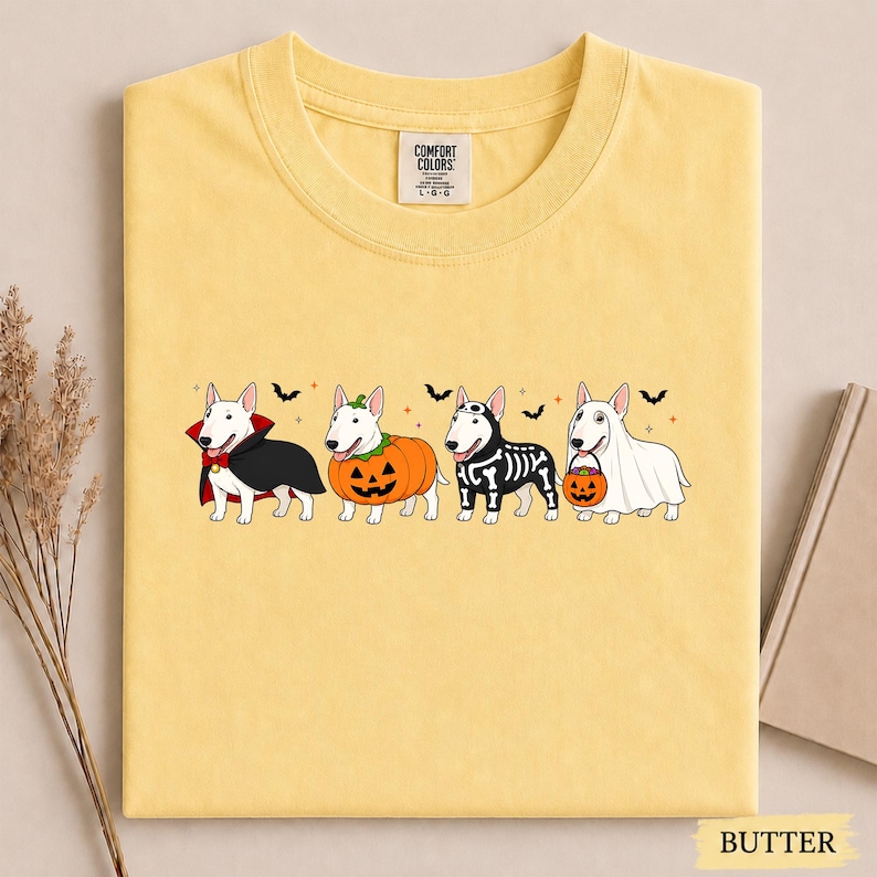 Puede incluir: Una camiseta color amarillo mantequilla con un gr&aacute;fico de cuatro perros Bull Terrier blancos con disfraces de Halloween: un vampiro, una calabaza, un esqueleto y un fantasma con un cubo de calabaza. La camiseta tiene una etiqueta Comfort Colors.