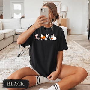 Puede incluir: Una camiseta negra de manga corta con un estampado gr&aacute;fico de cuatro perros bull terrier blancos vestidos con disfraces de Halloween, incluyendo un vampiro, una calabaza, un esqueleto y un fantasma, con peque&ntilde;os murci&eacute;lagos negros.