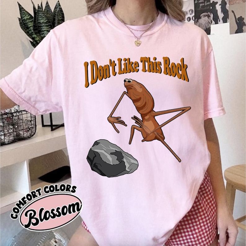 May include: A light pink t-shirt featuring a graphic of a brown insect-like creature standing near a grey rock. Above the creature, the text reads I Don't Like This Rock in brown letters. The shirt has short sleeves and a crew neckline.