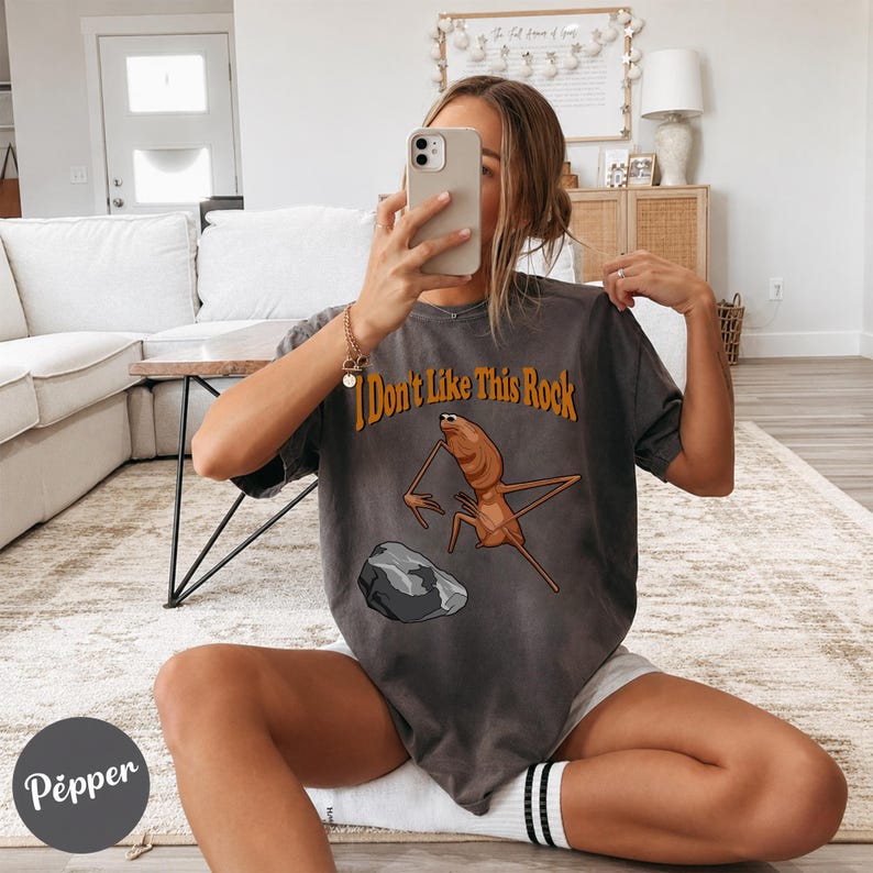 May include: A charcoal gray short-sleeve t-shirt featuring a graphic of a brown insect-like creature standing near a gray rock. The text "I Don't Like This Rock" is printed in orange above the creature. The shirt has a loose, relaxed fit.
