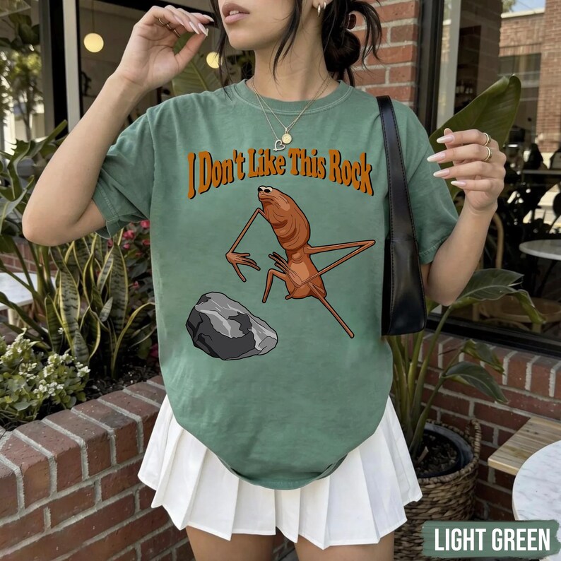 May include: A sage green short-sleeve t-shirt featuring a graphic of a brown insect-like creature standing near a gray rock. The text "I Don't Like This Rock" is printed in orange above the creature. The shirt has a crew neckline and a relaxed fit.