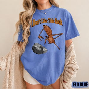 May include: A periwinkle blue t-shirt featuring a graphic of a brown, long-legged insect-like creature standing near a grey rock. Above the creature, the text reads "I Don't Like This Rock" in a bold, orange, serif font with a dark outline.