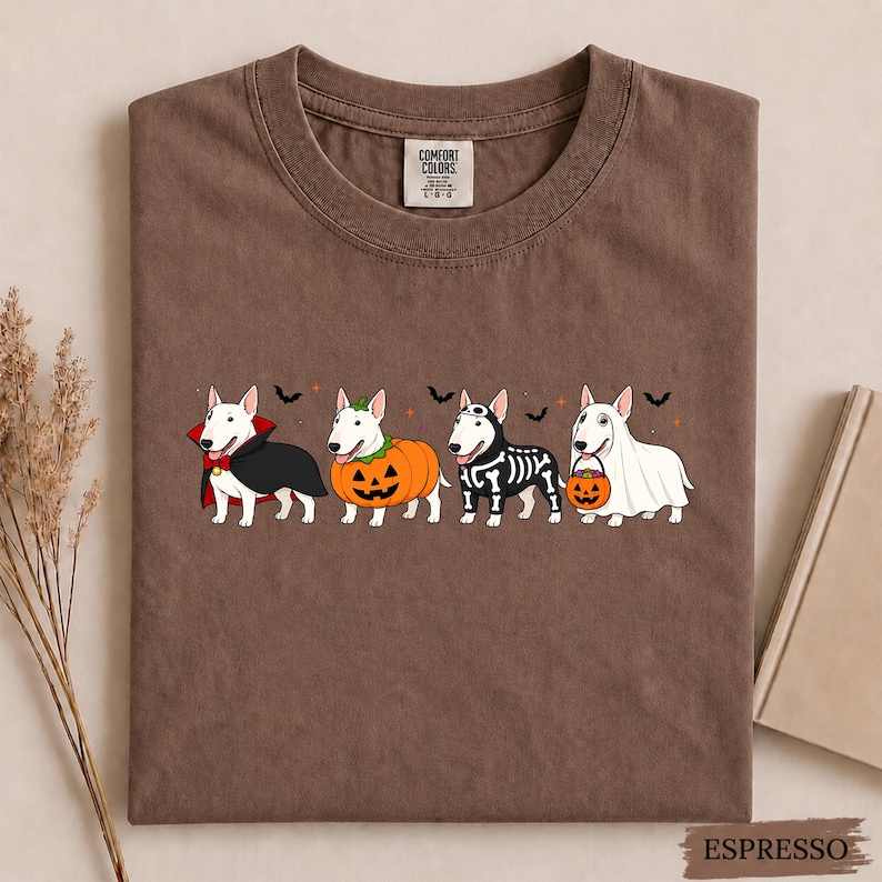 Puede incluir: Una camiseta marr&oacute;n con un gr&aacute;fico de cuatro perros Bull Terrier blancos disfrazados de Halloween: un vampiro con capa, una calabaza, un esqueleto y un fantasma con un cubo de dulces, junto a peque&ntilde;os murci&eacute;lagos negros y estrellas naranjas.