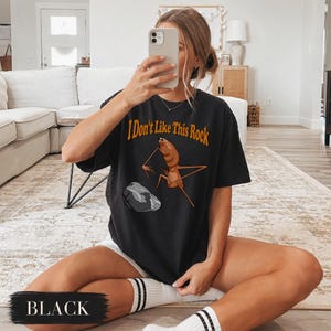 May include: A black short-sleeve t-shirt featuring a graphic of a brown insect-like creature standing near a grey rock. The text "I Don't Like This Rock" is printed in orange above the creature. The shirt has a crew neckline and a relaxed fit.
