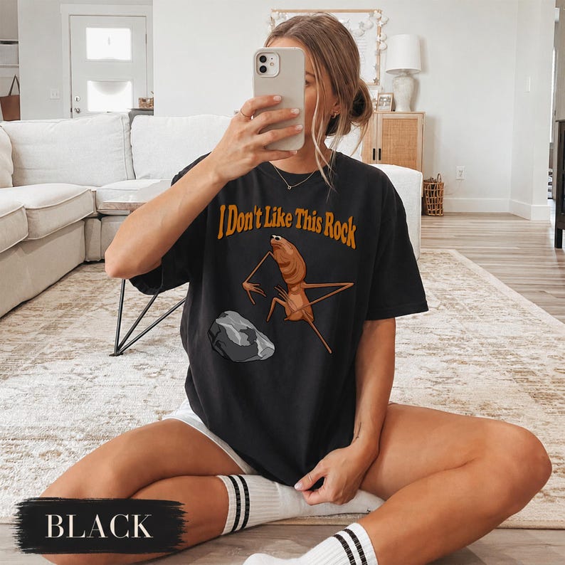 May include: A black short-sleeve t-shirt featuring a graphic of a brown insect-like creature standing near a grey rock. The text "I Don't Like This Rock" is printed in orange above the creature. The shirt has a crew neckline and a relaxed fit.