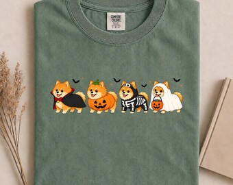 May include: A moss green crew neck t-shirt featuring a graphic of four Pomeranian dogs in Halloween costumes. The dogs are dressed as a vampire, a pumpkin, a skeleton, and a ghost holding a jack-o'-lantern bucket, with small bats and stars.