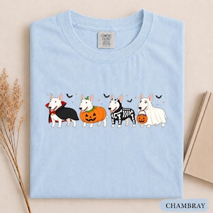 Puede incluir: Una camiseta azul claro con un gr&aacute;fico de cuatro perros bull terrier blancos con disfraces de Halloween: un vampiro, una calabaza, un esqueleto y un fantasma con un cubo de calabaza, decorado con peque&ntilde;os murci&eacute;lagos y estrellas.