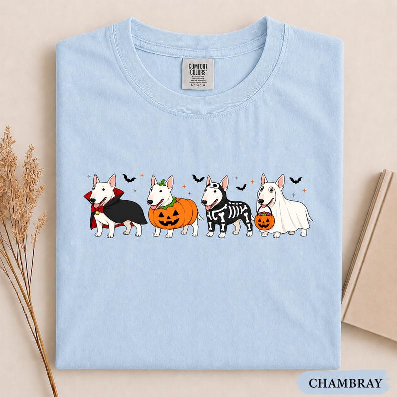 Puede incluir: Una camiseta azul claro con un gr&aacute;fico de cuatro perros bull terrier blancos con disfraces de Halloween: un vampiro, una calabaza, un esqueleto y un fantasma con un cubo de calabaza, decorado con peque&ntilde;os murci&eacute;lagos y estrellas.