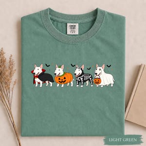 Puede incluir: Una camiseta verde salvia con un gr&aacute;fico de cuatro perros Bull Terrier blancos disfrazados de Halloween: un vampiro con capa, una calabaza, un esqueleto y un fantasma sosteniendo un cubo de calabaza, con peque&ntilde;os murci&eacute;lagos negros de fondo.