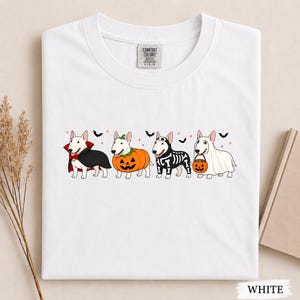 Puede incluir: Una camiseta blanca con un gr&aacute;fico de cuatro perros Bull Terrier con disfraces de Halloween. Los perros est&aacute;n vestidos de vampiro, calabaza, esqueleto y fantasma con un cubo de calabaza, decorado con peque&ntilde;os murci&eacute;lagos negros y estrellas.