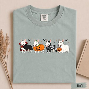 Puede incluir: Una camiseta verde salvia con un gr&aacute;fico de cuatro perros Bull Terrier blancos con disfraces de Halloween. Los perros est&aacute;n vestidos de vampiro, calabaza, esqueleto y fantasma con un cubo de dulces, decorado con peque&ntilde;os murci&eacute;lagos y estrellas.