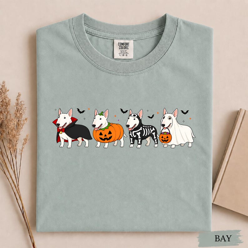 Puede incluir: Una camiseta verde salvia con un gr&aacute;fico de cuatro perros Bull Terrier blancos con disfraces de Halloween. Los perros est&aacute;n vestidos de vampiro, calabaza, esqueleto y fantasma con un cubo de dulces, decorado con peque&ntilde;os murci&eacute;lagos y estrellas.