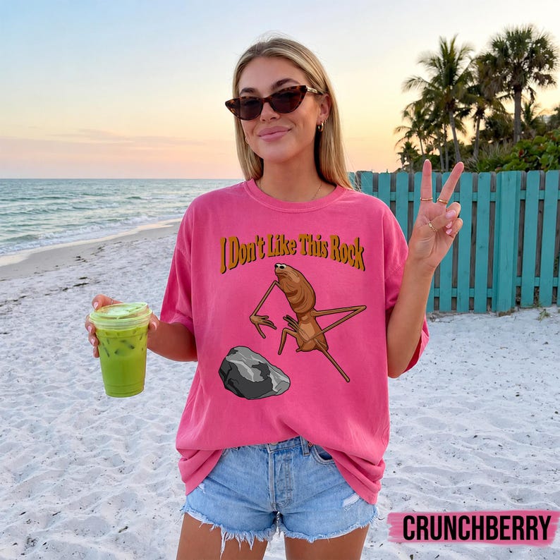 May include: A pink short-sleeve t-shirt featuring a graphic of a brown insect-like creature standing near a gray rock. The text "I Don't Like This Rock" is printed in a dark yellow font above the illustration. The shirt has a crew neckline.