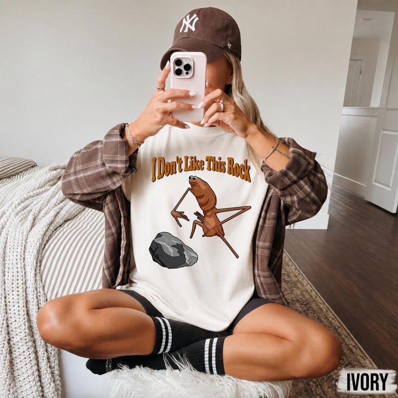 May include: An ivory t-shirt featuring a brown insect illustration and the text "I Don't Like This Rock" in brown letters above a grey rock. The shirt is worn with a brown plaid button-down shirt, a brown baseball cap, and black shorts.